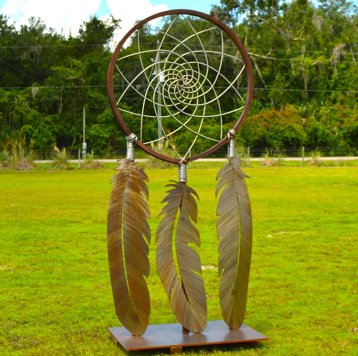 Full sculpture from another angle, showing the circular web and long feather forms against a blue sky.