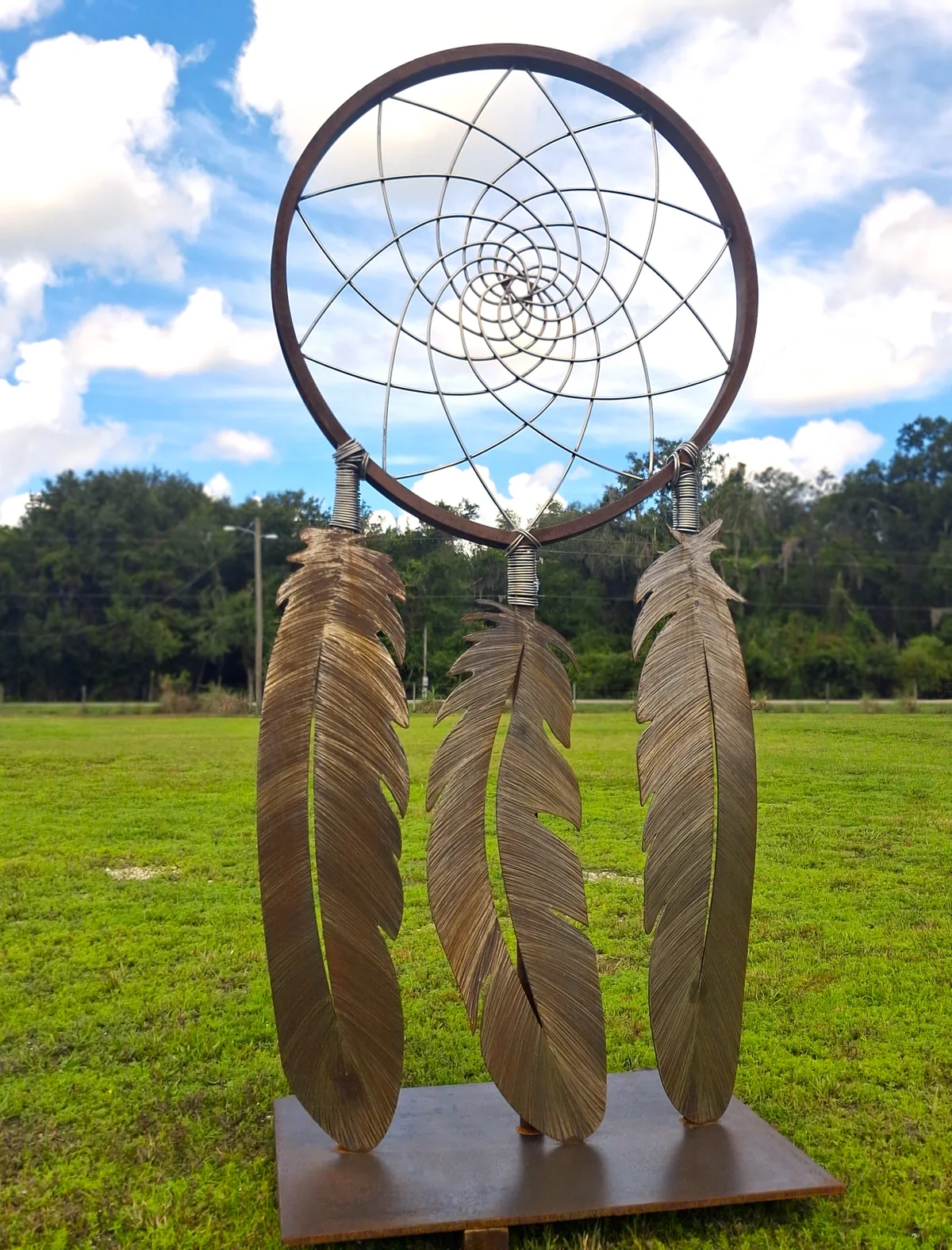 Tall steel sculpture with a circular web and three metal feathers arranged vertically, standing on a square base outdoors.