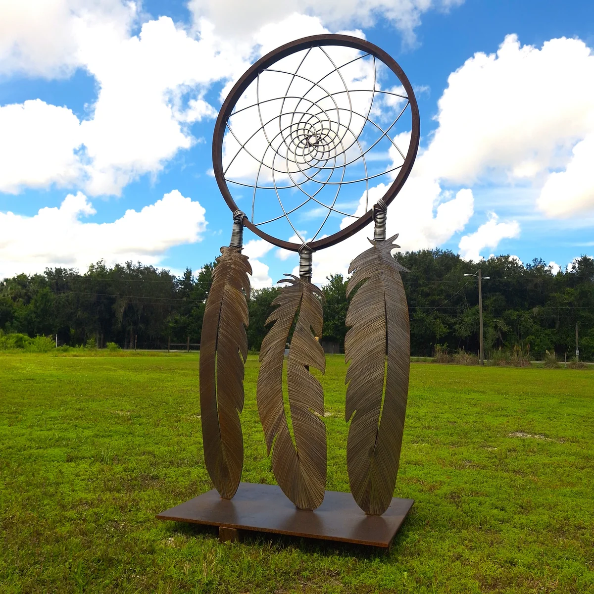 Full view of the circular web, three metal feathers, and square base in a grassy setting.
