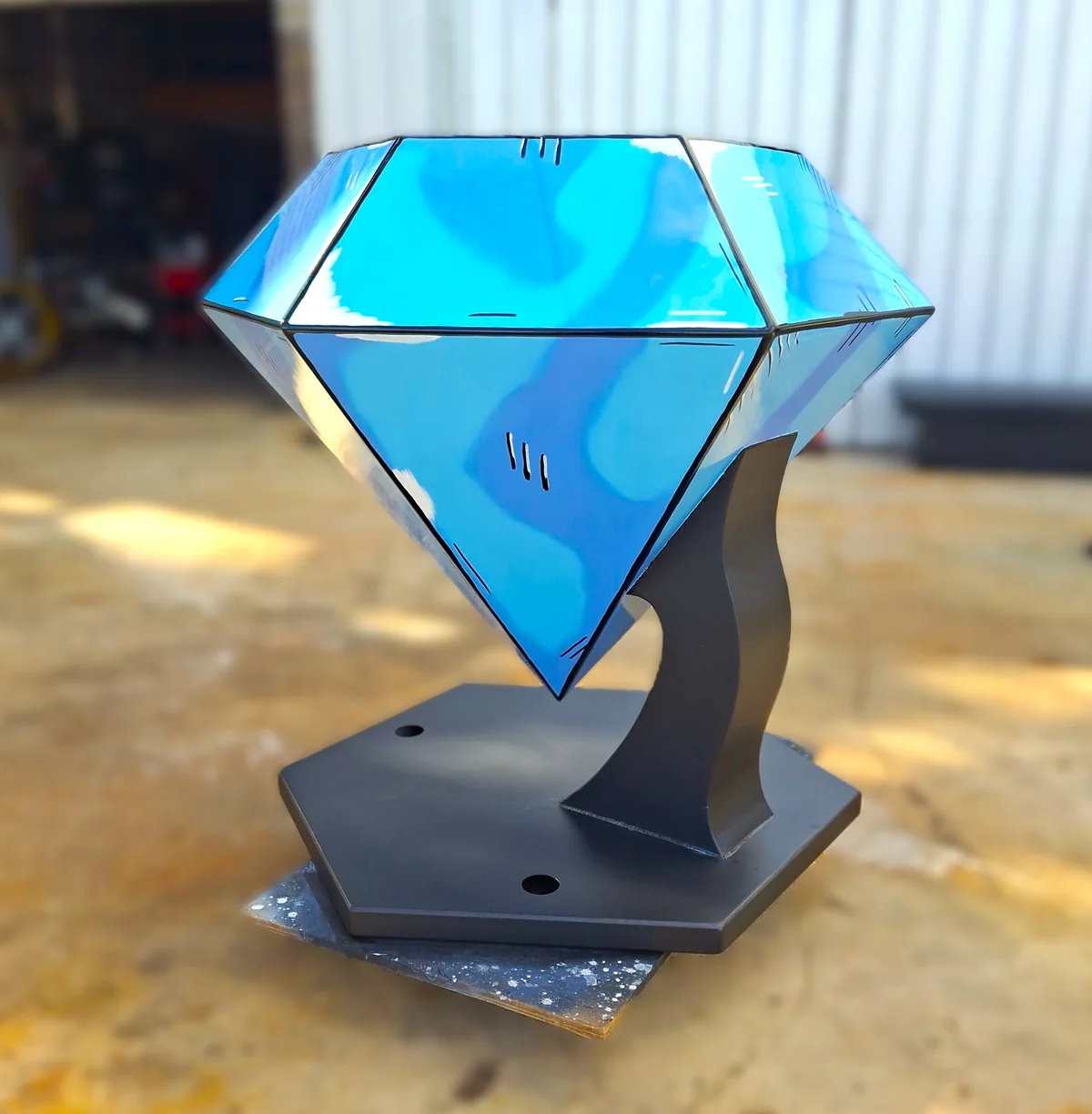 Blue faceted sculpture and curved black pedestal shown from another angle outdoors.