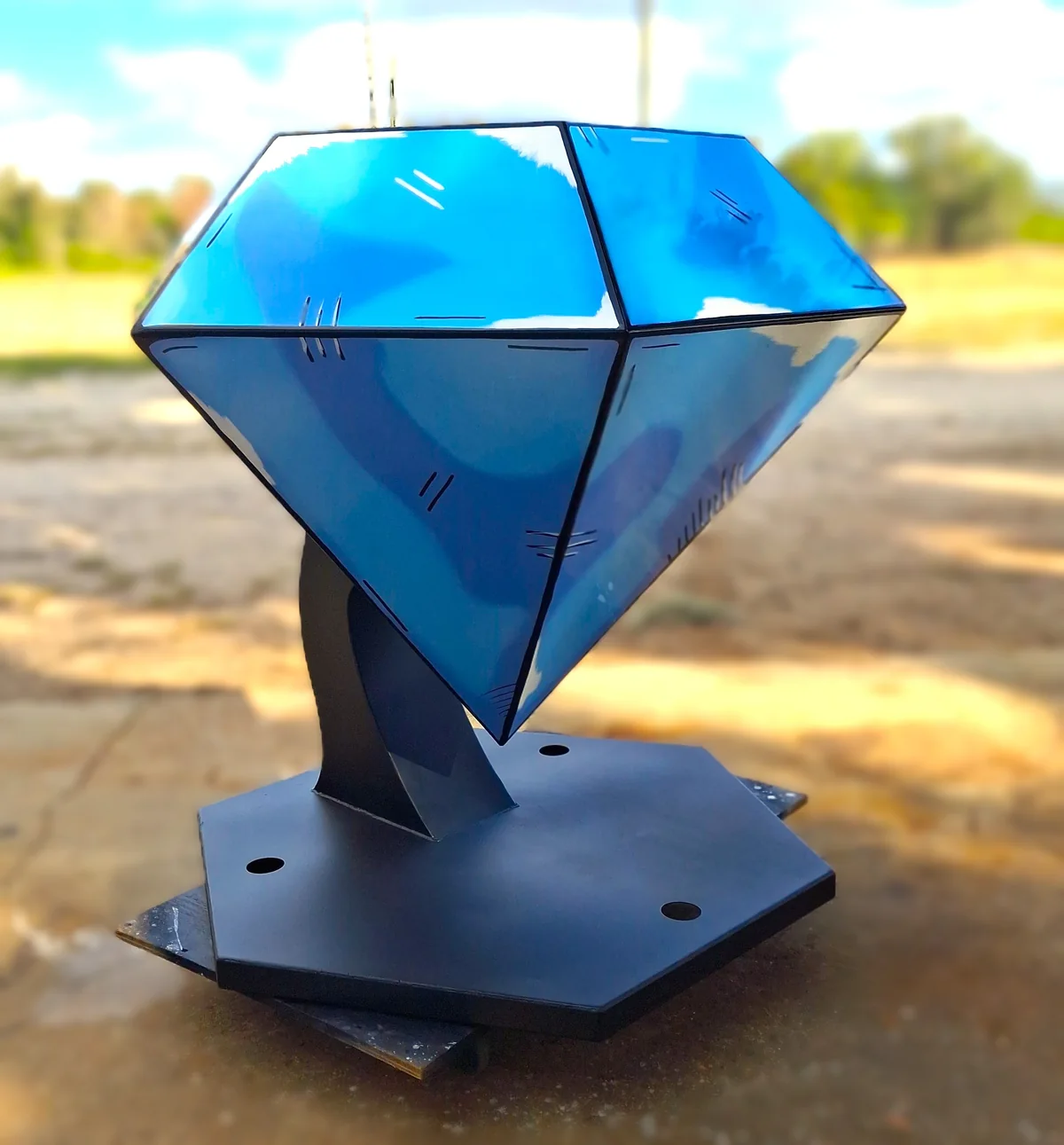 Blue faceted diamond-shaped sculpture on a black pedestal, viewed at an angle outdoors.