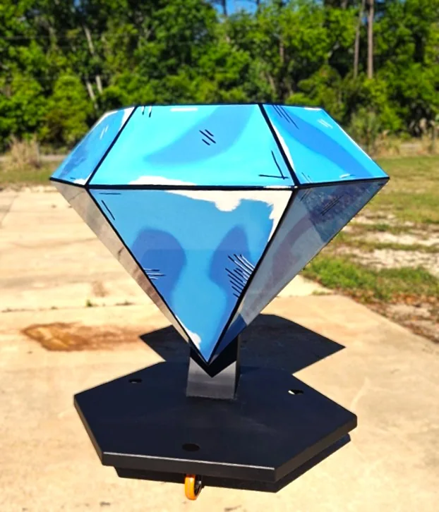 Front view of the blue diamond-shaped sculpture on a black pedestal, with trees in the background.